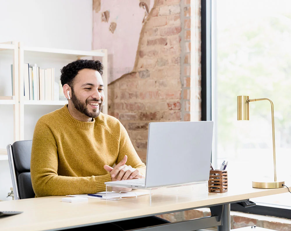 Man in a mustard yellow sweater smiling at his laptop, sitting down in his office