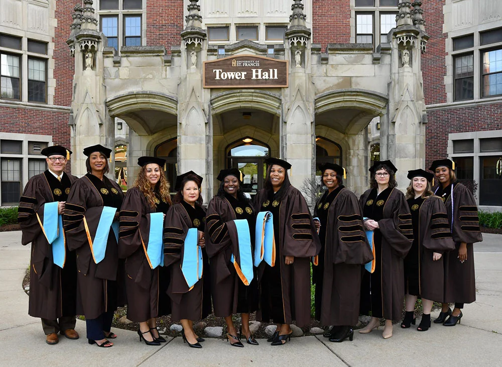 Group of grads wearing cap and gown outside of building on campus smiling