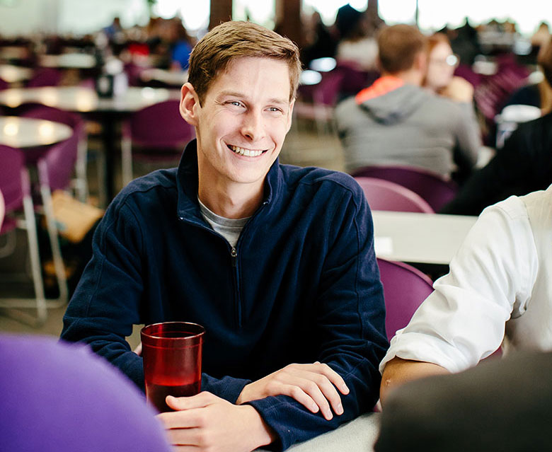 A man wearing a navy polo smiling in a cafeteria amongst conversation with others