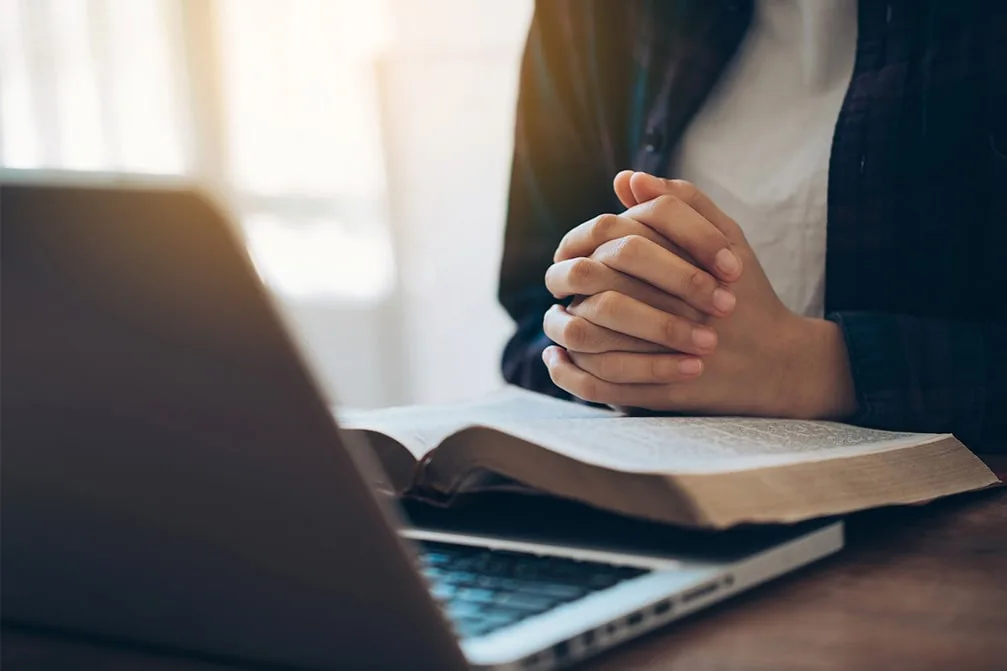 Hands folded on an open book beside a laptop